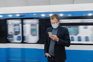 Horizontal shot of male worker poses at subway platform, commutes by public transport, uses modern mobile phone to check route, wears medical protective mask against coronavirus or flu. Health hazard photo