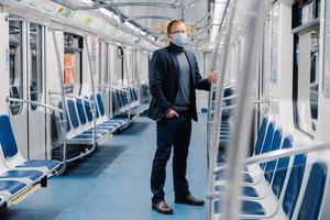 Full length shot of male entrepreneur in formal suit, medical mask, stands in urban train during disease outbreak, virus protection and quarantine protects himself from coronavirus at public transport photo