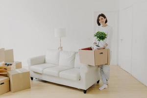Full length shot of cheerful brunette woman holds cardboard box with indoor plant, stands in spacious room with sofa and floor lamp, dressed in casual clothes. Female tenant stands in new home photo