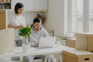 Indoor shot of happy woman and man spend morning at their new kitchen with big windows and modern furniture, husband works on laptop computer discusses with wife trendy design, surrounded with boxes photo