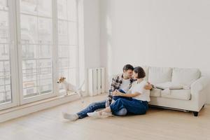 Horizontal view of happy affectionate family couple dressed in casual wear, embrace and express love to each other, pose on floor near sofa in modern apartment, their pet looks through big window photo