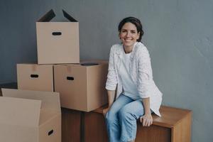 feliz adolescente se mueve. la señora hispana está sentada en cajas en un apartamento nuevo y sonriendo. foto