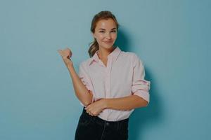 Attractive young female dressed in formal white shirt and black jeans indicates with thumb away photo