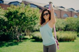 Beautiful glad young woman does arms exercises with fitness gum stands outdoor has athletic body wears sunglasses enjoys summer day has regular training. Sportswoman uses expander. Sport concept photo