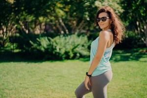 Pleased brunette European woman in active wear poses sideways wears sunglases smartwatch has fitness outdoors rests after jogging smiles pleasantly. Health body care sport and exercising concept photo