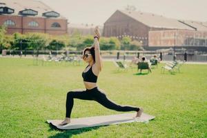 People sport and recreation concept. Sporty woman has healthy body raises arms exercises on karemat outdoors wears cropped top and leggings enjoys workout outside. Sportswoman in good shape. photo