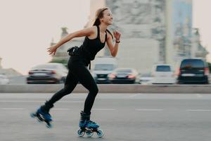 joven mujer sana delgada de cabello oscuro tiene tiempo de recreación disfruta patinar en velocidad se mueve rápido tiene expresión alegre usa camiseta negra y polainas. descanso activo y concepto de entrenamiento deportivo foto