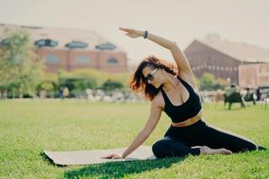 Horizontal shot of motivated young female model has regular morning workout raises arm and leans at different sides dressed in sportswear poses on karemat outside stays in good physical shape photo