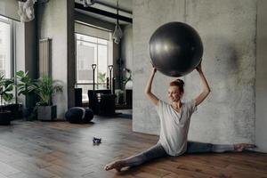 hermosa mujer fitness en posición dividida sosteniendo una pelota de ejercicio sobre la cabeza y disfrutando del entrenamiento de pilates foto