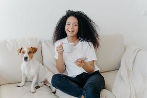 Delighted dark skinned woman poses in modern apartment, sits on comfortable sofa with pet, drinks coffee, uses mobile phone for online communication, being in good mood, flipping news, uses app