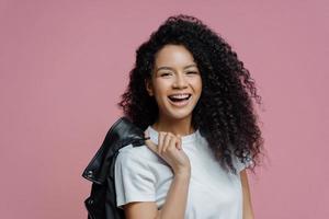 Portrait of positive African American woman smiles toothily, being in good mood after walk in park, dressed in white t shirt holds leather jacket on shoulder isolated on pink background. People, style