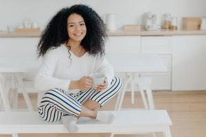 Happy morning concept. Photo of cheerful curly African American woman sits in lotus pose at white bench, sips tasty aromatic drink, feels relaxed, poses against kitchen interior, smiles broadly