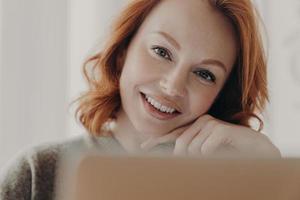 Cropped shot of cheerful pleasant looking ginger European woman with white teeth poses near modern laptop computer, uses internet connection for freelance, being in good mood, earns money online