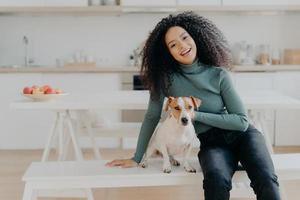Female pet owner expresses love to dog, dressed in casual turtleneck and jeans, sits at white bench against cozy kitchen interior, spends free time at home, smiles broadly from happy enotions photo