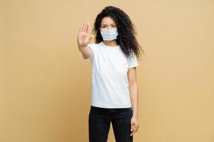 Say no to spreading disease. Serious looking Afro American woman makes stop gesture, wears medical mask, prevents virus Covid-19, stands indoor, isolated. Stop infectious disease immediately photo