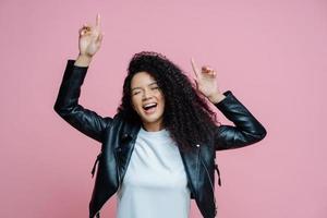 Carefree beautiful Afro American woman dances with arms raised, moves with rhythm of music on disco party, wears white t shirt and leather jacket, closes eyes with pleasure, keeps mouth opened photo