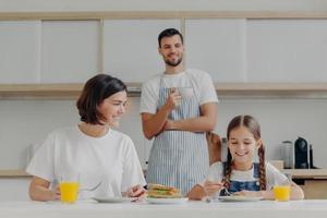 Happy mother talks to daughter while have breakfast. Father stands behind, prepared delicious dish for family. Friendly family members meet at kitchen during weekend, enjoy nice conversation photo