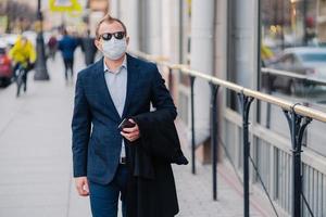 Outdoor shot of businessman in formal clothes, holds modern cell phone in hands, walks on street, protects himself with medical mask, prevents transmissible infectious disease or air pollution in city photo