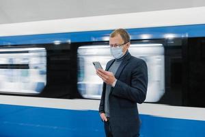 Horizontal shot of male traveler uses public transport for commuting, wears medical mask to protect from coronavirus or covid-19, waits for train, uses mobile phone, sends text messages in chat photo