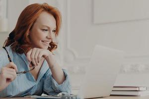 Horizontal shot of redhead European woman poses at home office, focused at laptop screen, holds spectacles, watches video on computer, enjoys good news, wears shirt, enjoys distance learning photo