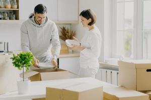 Photo of glad married woman and man unpack dinnerware from boxes, dressed casually, hold plates, pose in new modern kitchen, surrounded with moving packages. People, home and relocation concept