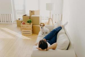 Indoor shot of relaxed woman rests on sofa with favourite pet, removes in new rented apartment, pile of carton boxes and lamp near. Female homeowner feels happy of being in new home. Relocation