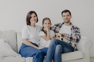 Photo of happy friendly family of mum, dad and daughter pose on comfortable sofa with pet, have good relationship, dressed casually, smile positively. Family time and domestic atmosphere concept