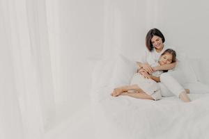Photo of relaxed young mother embraces daughter with love, pose in pyjamas bare foot, have pleasant smiles, being in spacious white bedroom, have good relationships, wake up late in morning.