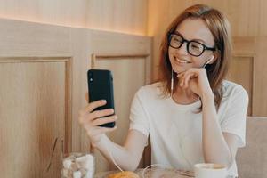 Smiling happy woman in spectacles wears earphones uses smartphone in cafe photo