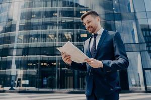 propietario de un negocio sonriente leyendo el periódico mientras camina cerca del edificio de oficinas foto