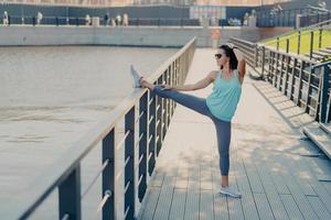 mujer joven en forma con cabello oscuro estira las piernas en la valla se calienta antes de trotar usa gafas de sol camiseta polainas y zapatillas de deporte se prepara para el entrenamiento cardiovascular plantea al aire libre estar en buena forma física foto