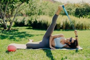 Active woman does workout outside. Slim brunette woman stretches legs with sports gum dressed in activewear lying on fitness mat against green nature background has good flexibility. Exercising photo