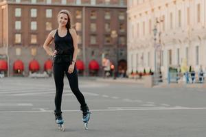 concepto de deporte y recreación. la toma horizontal de una mujer activa practica poses de patinaje sobre cuchillas mantiene la mano en la cintura el fondo borroso de la ciudad toma un descanso. patinar después del trabajo. vida saludable foto