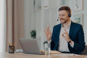 Career and distance job concept. Pleased bearded man corporate worker dressed formally gestures okay sign at webcam of laptop uses headset for online calling poses at desktop gains new knowledge