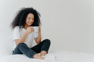 African American woman sips hot drink, poses in bed, enjoys good morning, dressed casually, spends weekend at home, blank space aside for your advertising. People, coziness and rest concept.