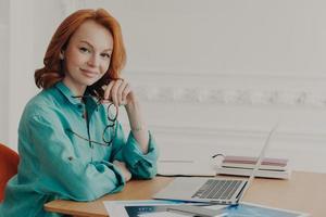 Smiling pleased young red head woman ready for brainstroming with colleagues online, poses in coworking space with paper documents, going to participate in online conference, works from home photo