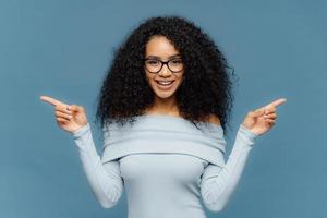 Happy woman with Afro hairstyle, points sideways with both index fingers, wears casual jumper, hesitates between two objects, isolated over blue background. Female indicates left and right indoor photo