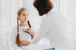 Caring female pediatrician listens childs lungs or heart with phonendoscope. Little girl comes on examination in private clinic, has every year doctor visit, explains symptoms of her disease