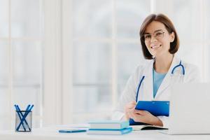 Female doctor writes prescription on special form, works in private clinic, wears white medical gown, ready for seeing patients, poses at workplace. Smiling physician or medical worker holds clipboard photo