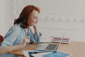 Profile shot of pleased redhead woman entrepreneur enjoys video conference with colleague, poses at desktop with paper documents, pleased to work from home, glad to see friend. Vitrual reality photo