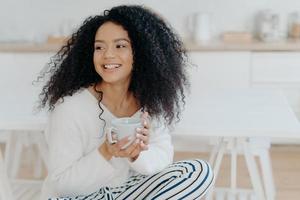Photo of cute cheerful woman with bushy curly hair looks away with smile, holds mug of coffee, wears casual clothing, enjoys cozy calm morning, savors taste. Afro lay in kitchen during day off