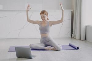 Slim sporty girl stretching on mat. Online remote fitness classes student watching video on laptop. photo