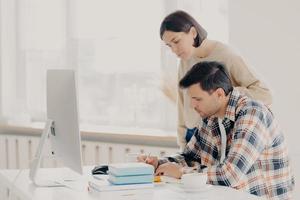 Husband and wife work on paper report together, check information, sit in front of computer, drinks coffee, dressed in casual wear, collaborate for teamworking. Family and remote work concept photo