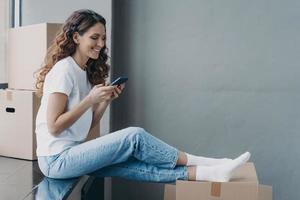 Happy attractive hispanic woman is sitting on the sill with boxes and texting on mobile phone. photo