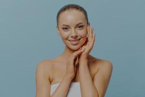 Young pretty woman with soft and healthy skin touching cheek, standing half-naked on blue background photo