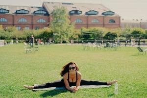 Fit active European woman does pilates on fitness mat wears sunglasses and sportswear shows great progress poses on green lawn has happy expression trains outside. Fitness and stretching concept photo