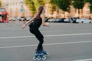 Active lifestyle and hobby concept. Sporty young woman does sport outdoors rides rollerblading dressed in sportsclothes enjoys fitness activities poses in urban place on road. Rollerskating.