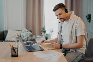 Focused bearded guy using laptop and notebook while working remotely photo