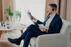 Sideways shot of bearded male corporate worker analyzes data looks through documents with graphics reviews financial report dressed in formal clothes poses in modern apartment uses laptop headset