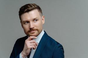 Thoughtful bearded businessman holds chin considers something looks away makes decision dressed in formal clothes looks forward future opportunity isolated over grey background blank space for text photo